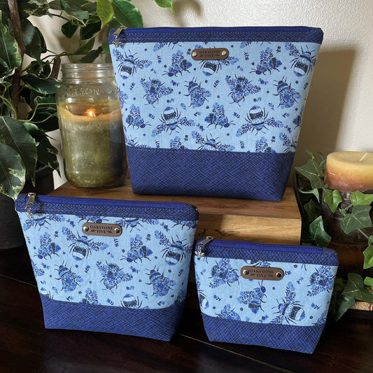 Set of 3 blue floral bumble bee patterned zipper pouches, sitting on a wooden table with a candle and plants.