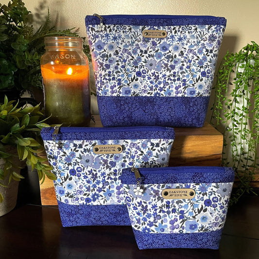 Set of three blue floral-patterned zipper pouches with "Oakstone Five" branding on a wooden surface with plants and a candle in the background.
