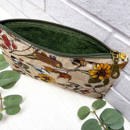 A zipper pouch with a beige floral and bird print exterior, a dark green vines interior lining, and a brass hardware, styled on a white surface with green leaves and a white brick wall background.