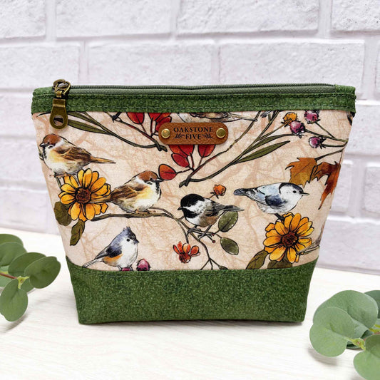 A medium sized zippered pouch with a green vines fabric trim and base, featuring a central printed panel of birds and flowers on a beige background, with a brown faux leather Oakstone Five brand patch on the front.