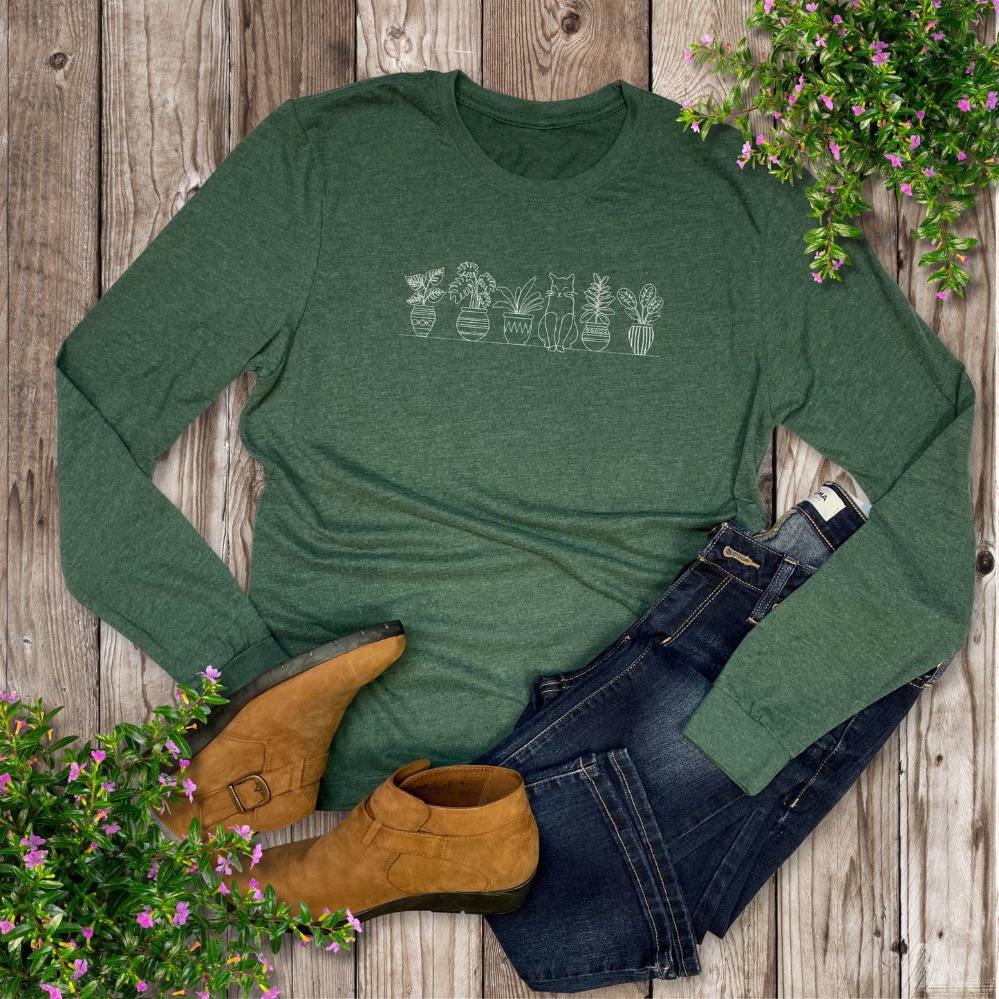Green long-sleeve shirt with cat sitting with potted plants design, paired with jeans and boots on a wooden surface.