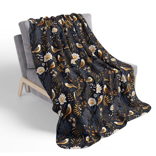 A nature-inspired sherpa blanket featuring a dark background with a bird and botanical motif, laying draped across a chair, on a white background