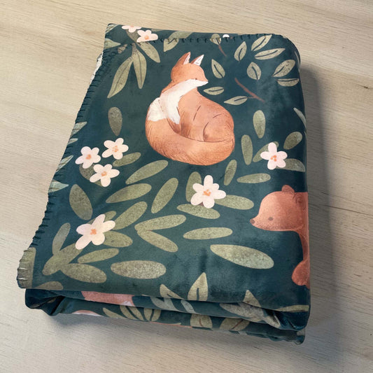 A folded sherpa throw blanket with forest-themed pattern featuring forest animals, laying on a wooden table