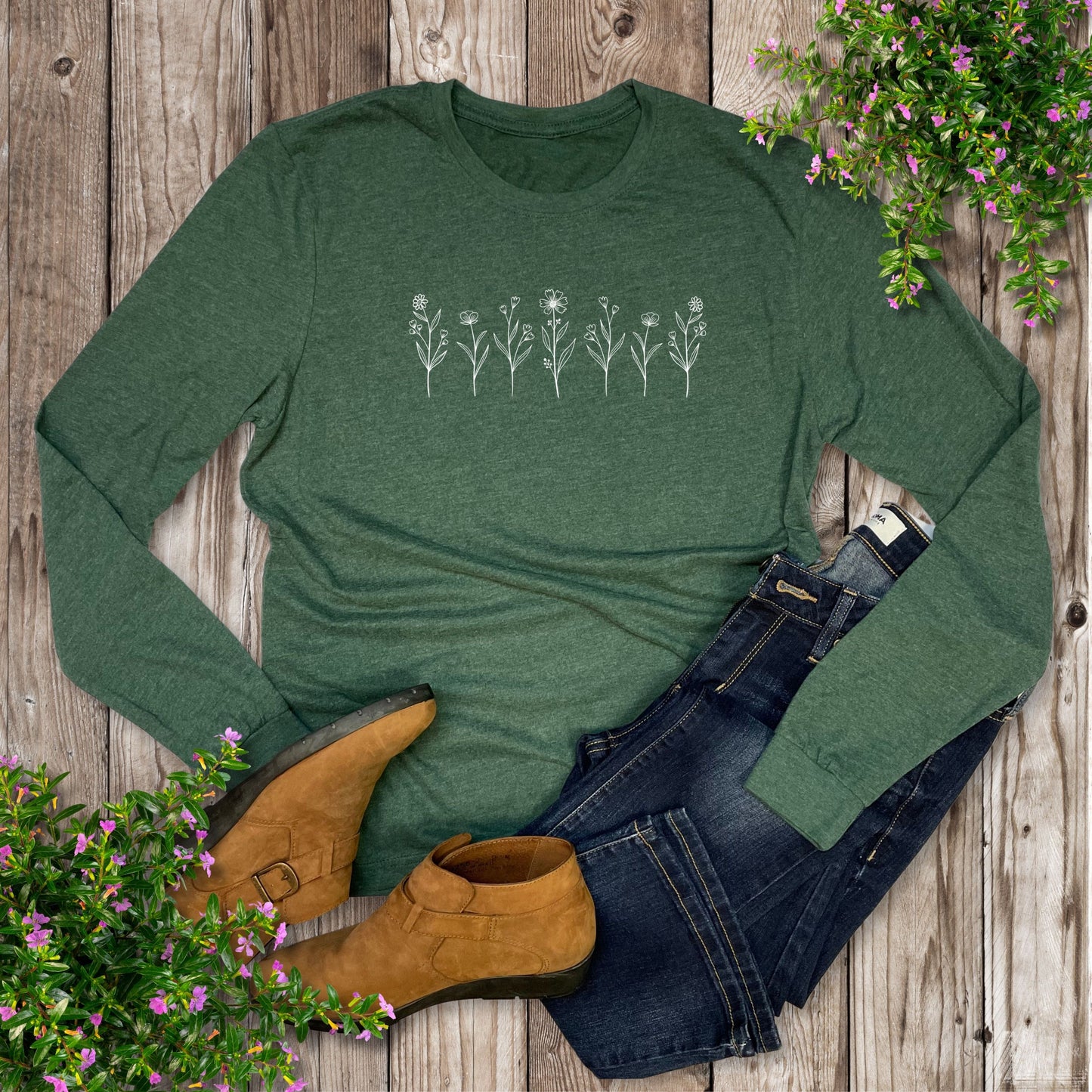 A green long sleeve shirt with a design of a row of mini white wildflowers on the front, laying on a wood table with plants, shoes, and jeans.