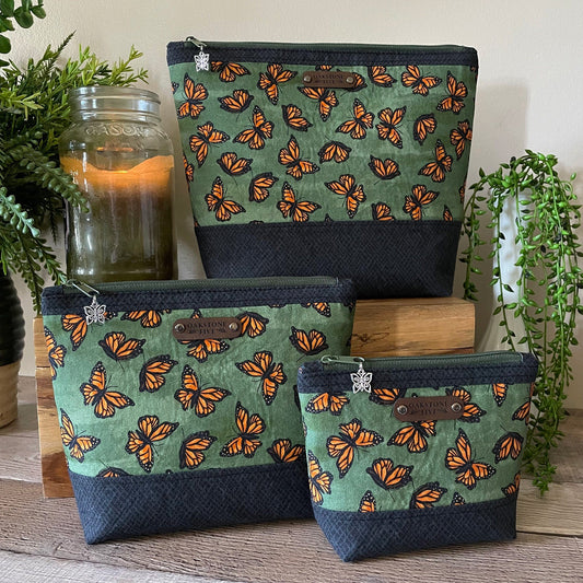 Three green zipper pouches with orange monarch butterfly pattern on a wooden surface with plants and a candle in the background.