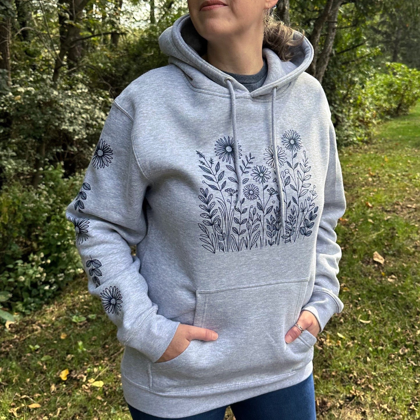 A woman standing outside in the woods wearing a fun, light grey hoodie that navy wildflowers on the front and on both sleeves.