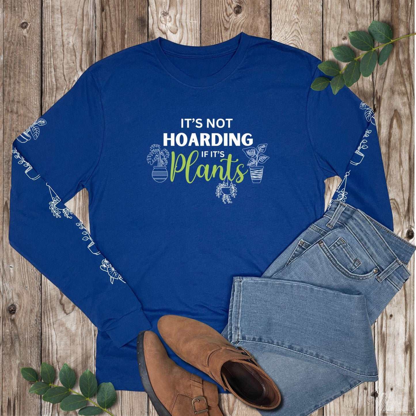 Blue long-sleeve shirt with plant-themed text that says "it's not hoarding if it's plants" and has potted plants down both sleeves, laying on a wooden surface with jeans and boots.