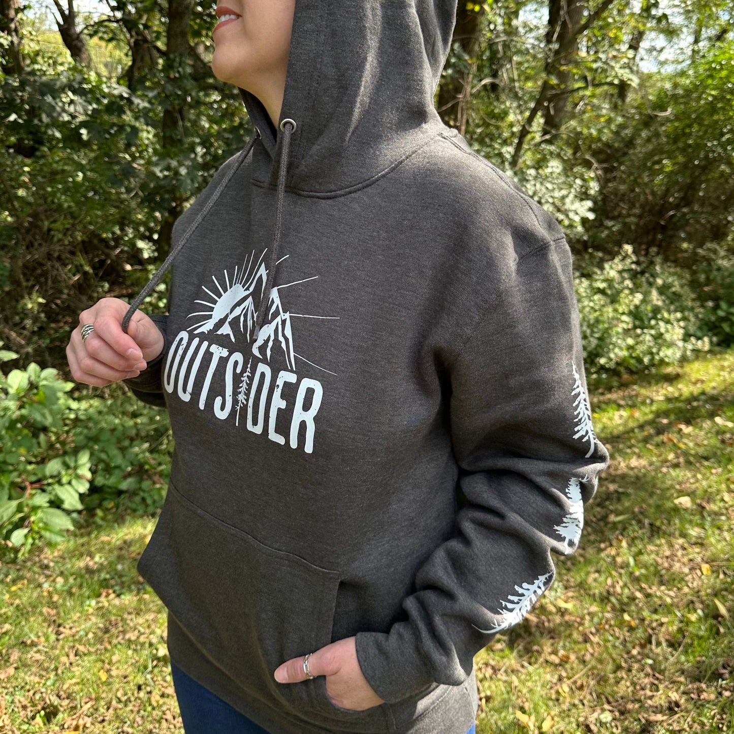 Woman wearing a gray hoodie that says "Outsider" on the front and has a sun and mountain scene, and trees going down the sleeves.