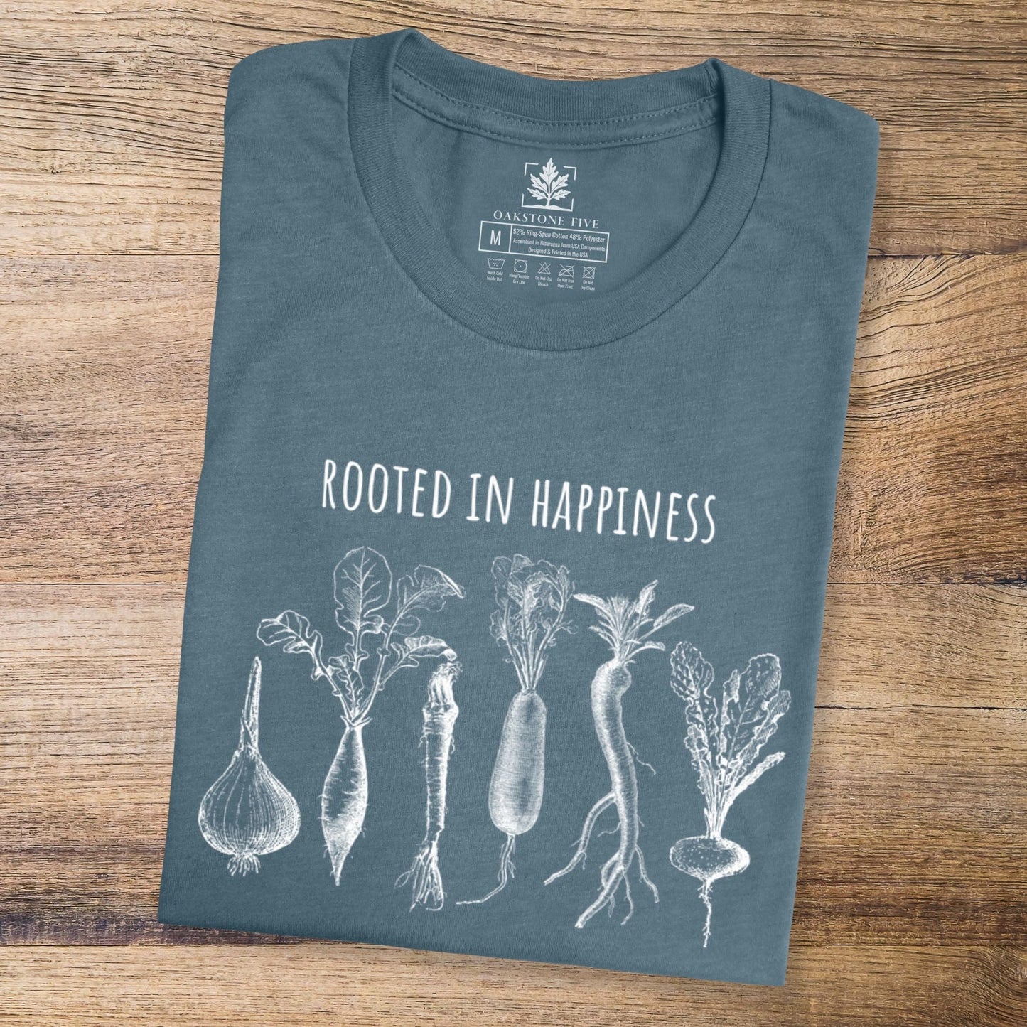 A folded slate blue positivity shirt for gardeners and plant lovers that has vegetables on it and says, "Rooted in Happiness".