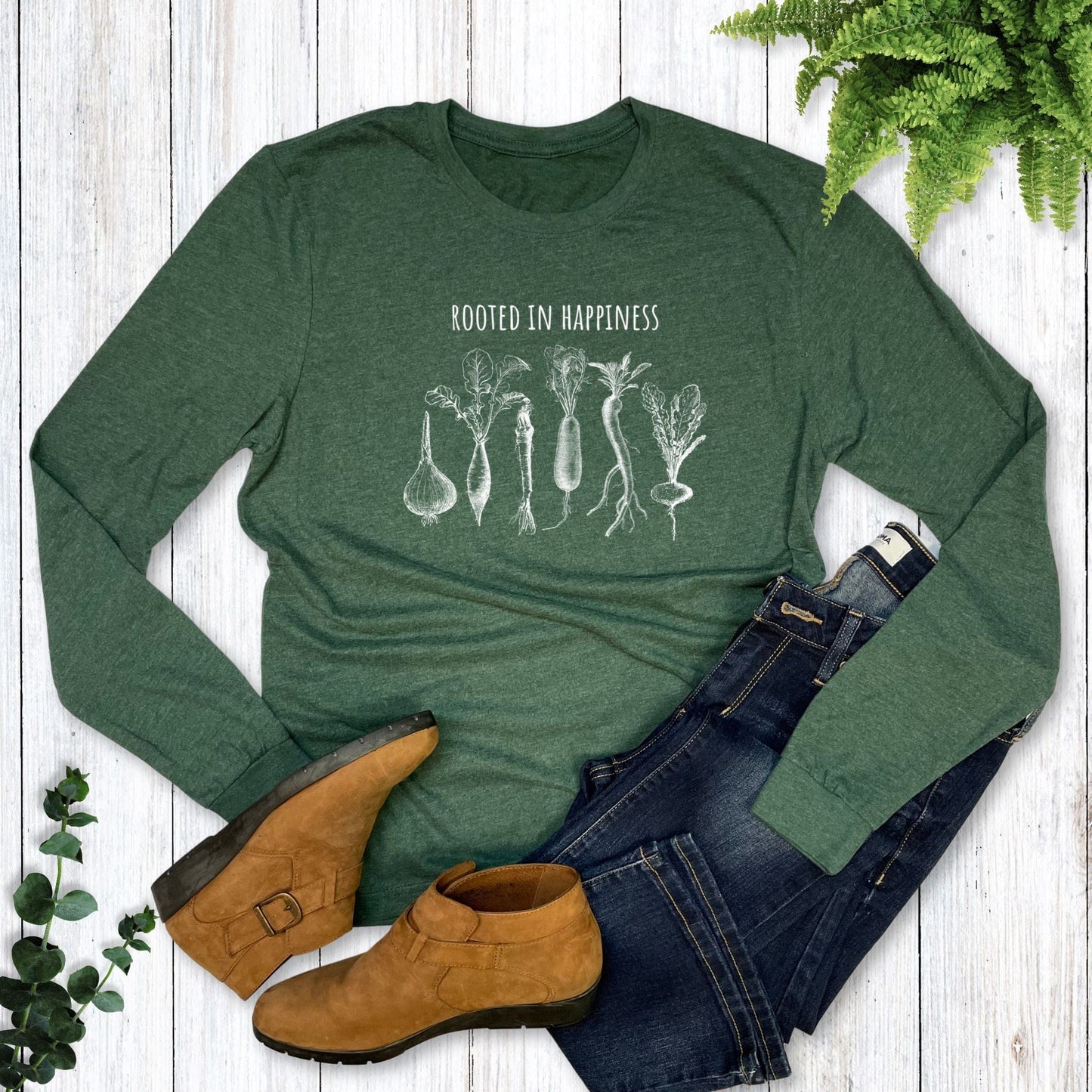 Green long-sleeve shirt with 'Rooted in Happiness' text and root vegetable illustrations on a wooden surface with jeans and boots.