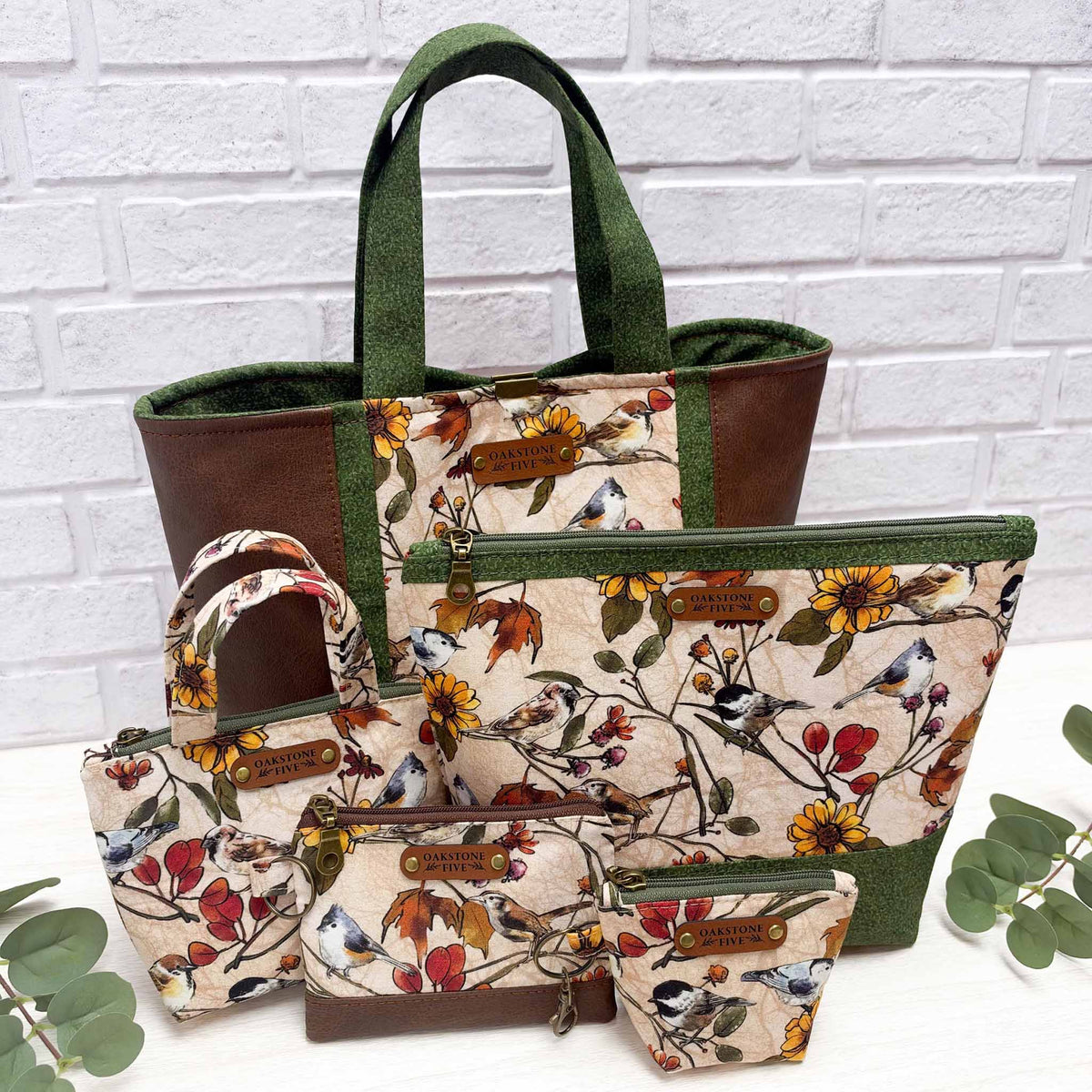 A handcrafted collection of floral bird patterned tote bags, zipper pouches, and little accessories sitting in a group with a white brick background and white table with greenery leaves.