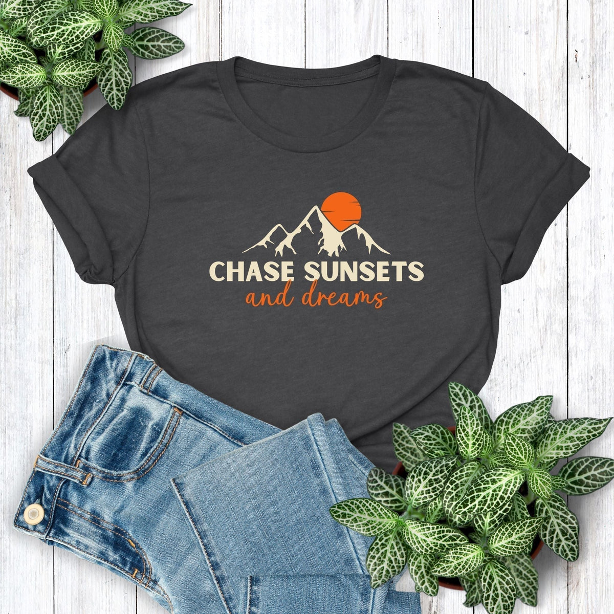 A fun, positive, nature t-shirt for sunset chasers that has an image of mountains and a sun and says, Chase Sunsets and Dreams.