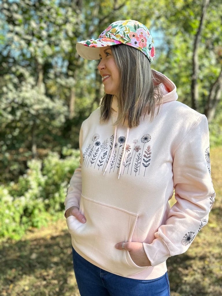 Woman standing in the woods, wearing a fun floral baseball hat and a pink hoodie with a flower design on the front and down both sleeves