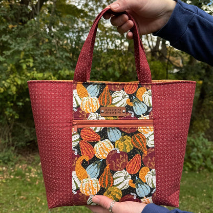 Woman outdoors among trees, holding a seasonal tote bag with a Fall gourd and pumpkin pattern.