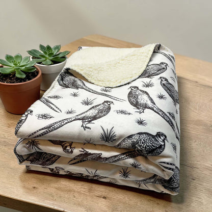 Folded sherpa blanket with pheasant bird pattern on a wooden surface with plants.