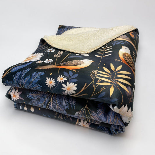 A folded sherpa blanket featuring a dark background with a bird and botanical motif, on a white background