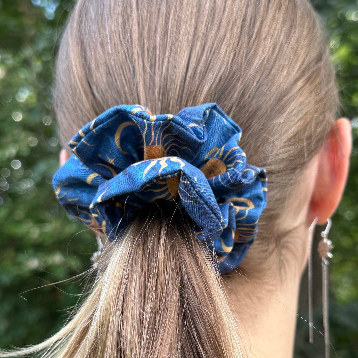 An outdoor photo of a woman wearing a blue sun, stars, and moon scrunchie to hold her ponytail.