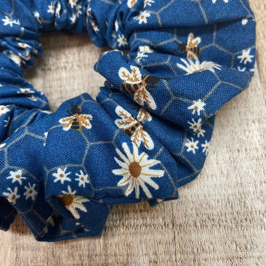 Blue floral scrunchie that has a daisies and honey bees pattern, laying on a wooden surface