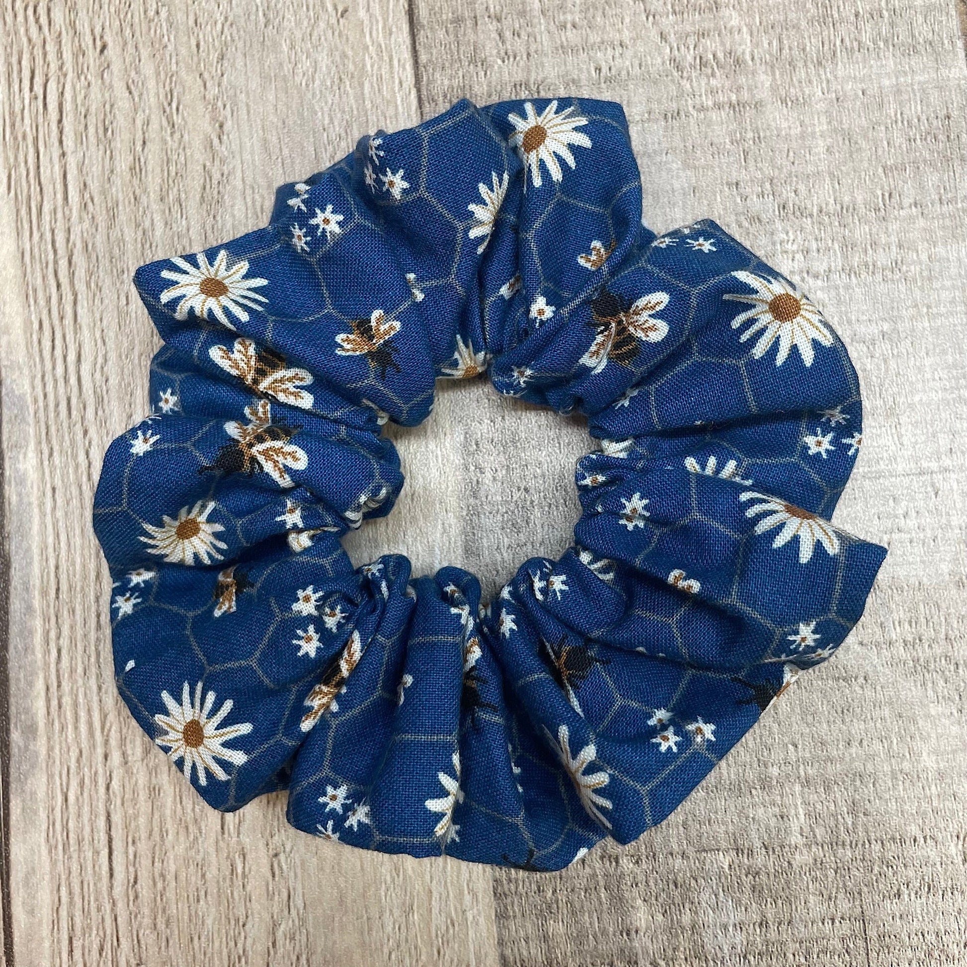 Blue floral scrunchie that has daisies and honey bees on a honeycomb pattern laying on a wooden surface