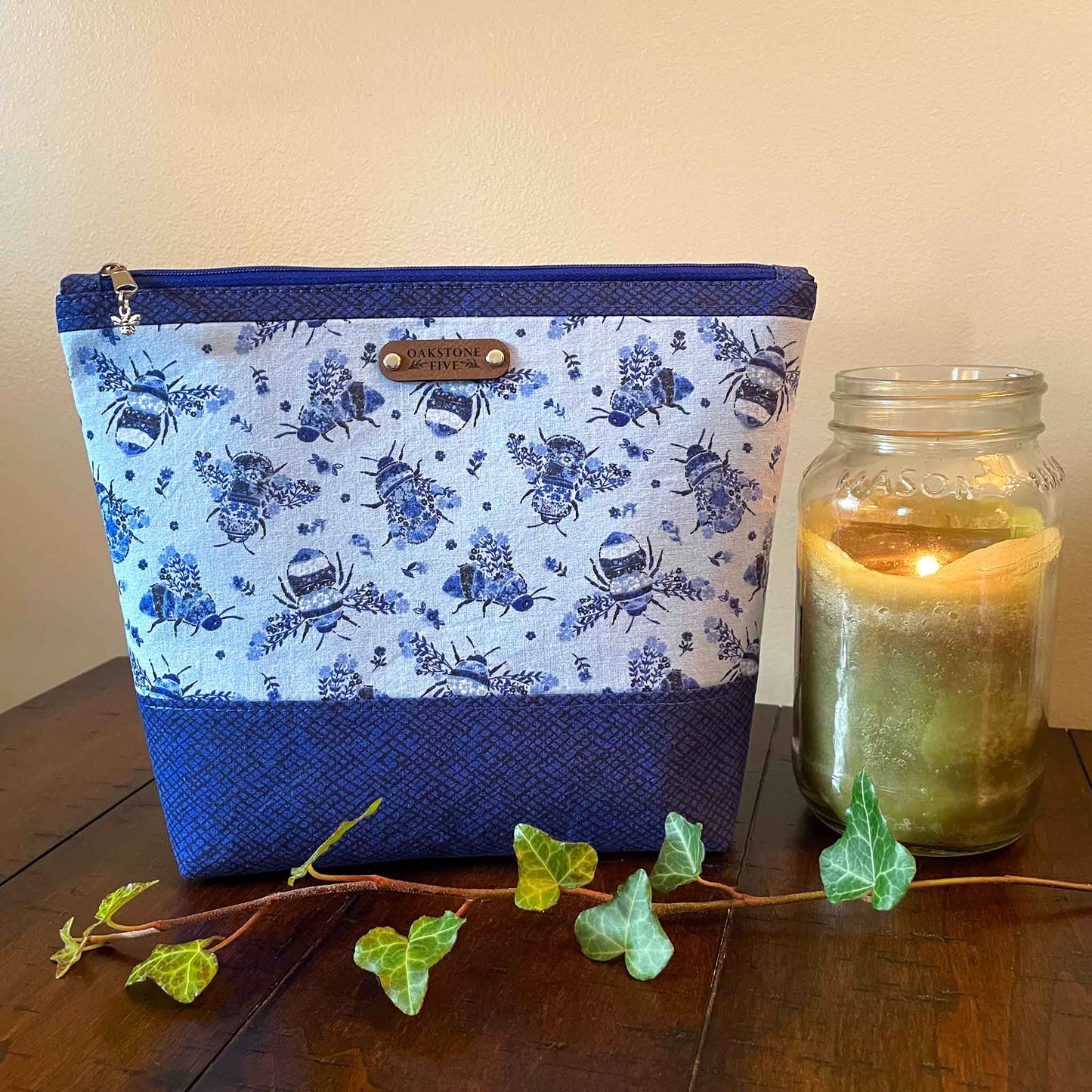 Large blue floral bumble bee patterned zipper pouch with a lit jar candle and ivy, sitting on a wooden table 