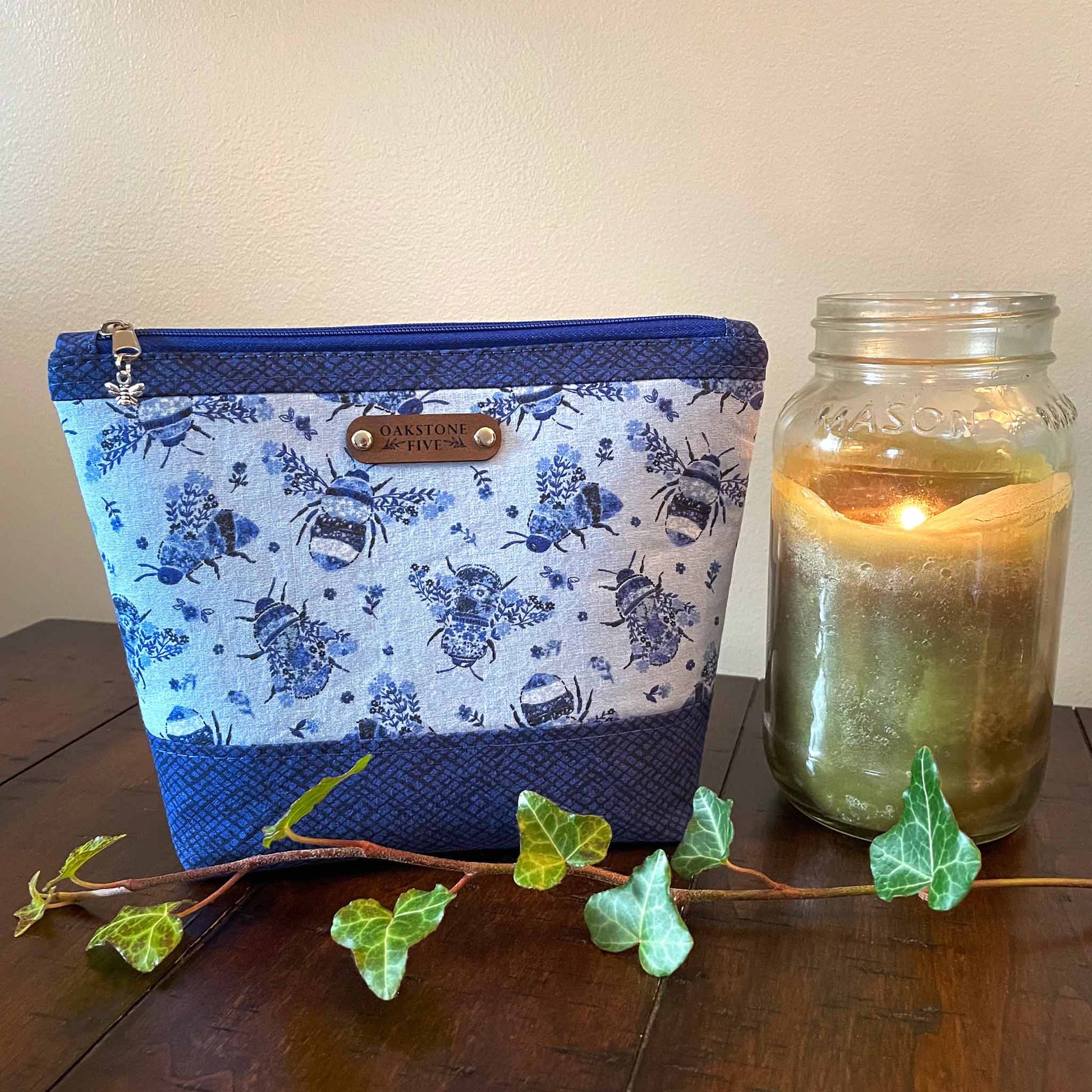 Medium blue floral bumble bee patterned zipper pouch with a lit jar candle and ivy, sitting on a wooden table 
