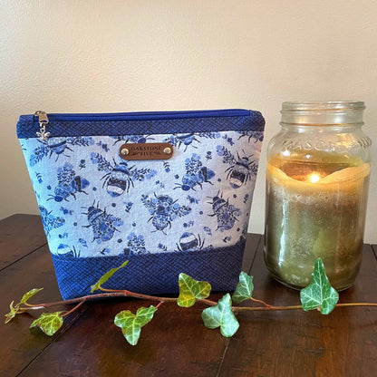 Medium blue floral bumble bee patterned zipper pouch with a lit jar candle and ivy, sitting on a wooden table 