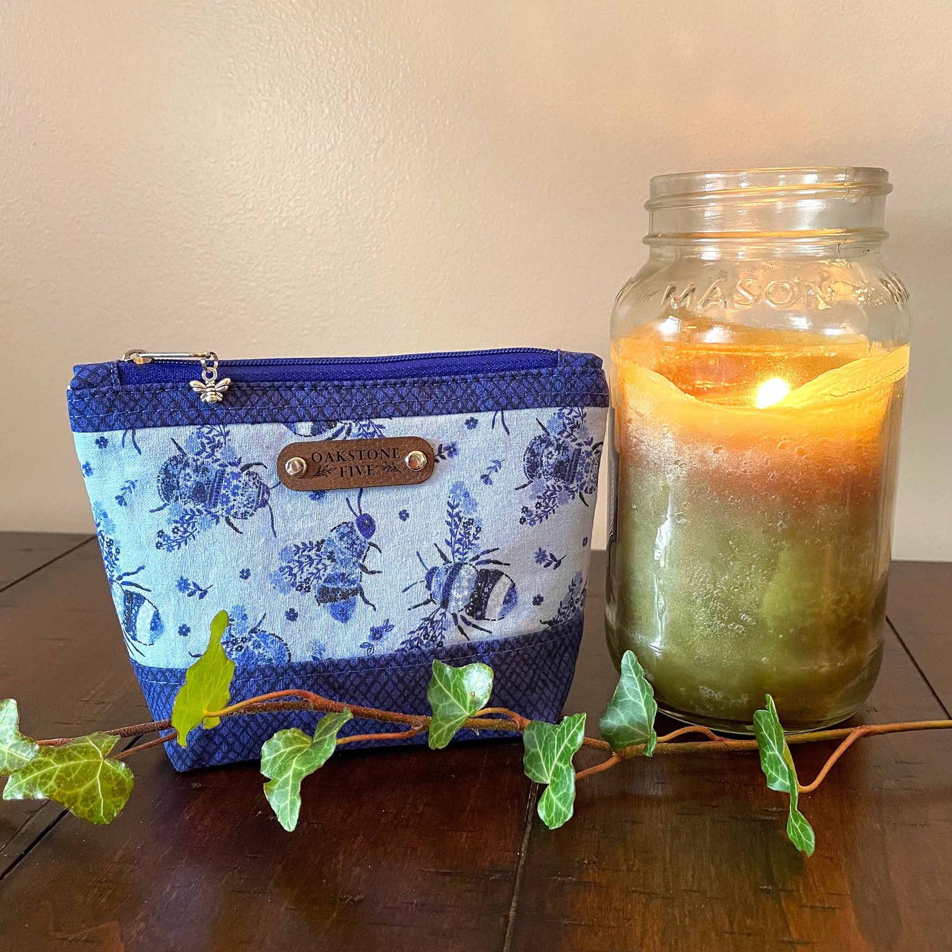 Small blue floral bumble bee patterned zipper pouch with a lit jar candle and ivy, sitting on a wooden table 