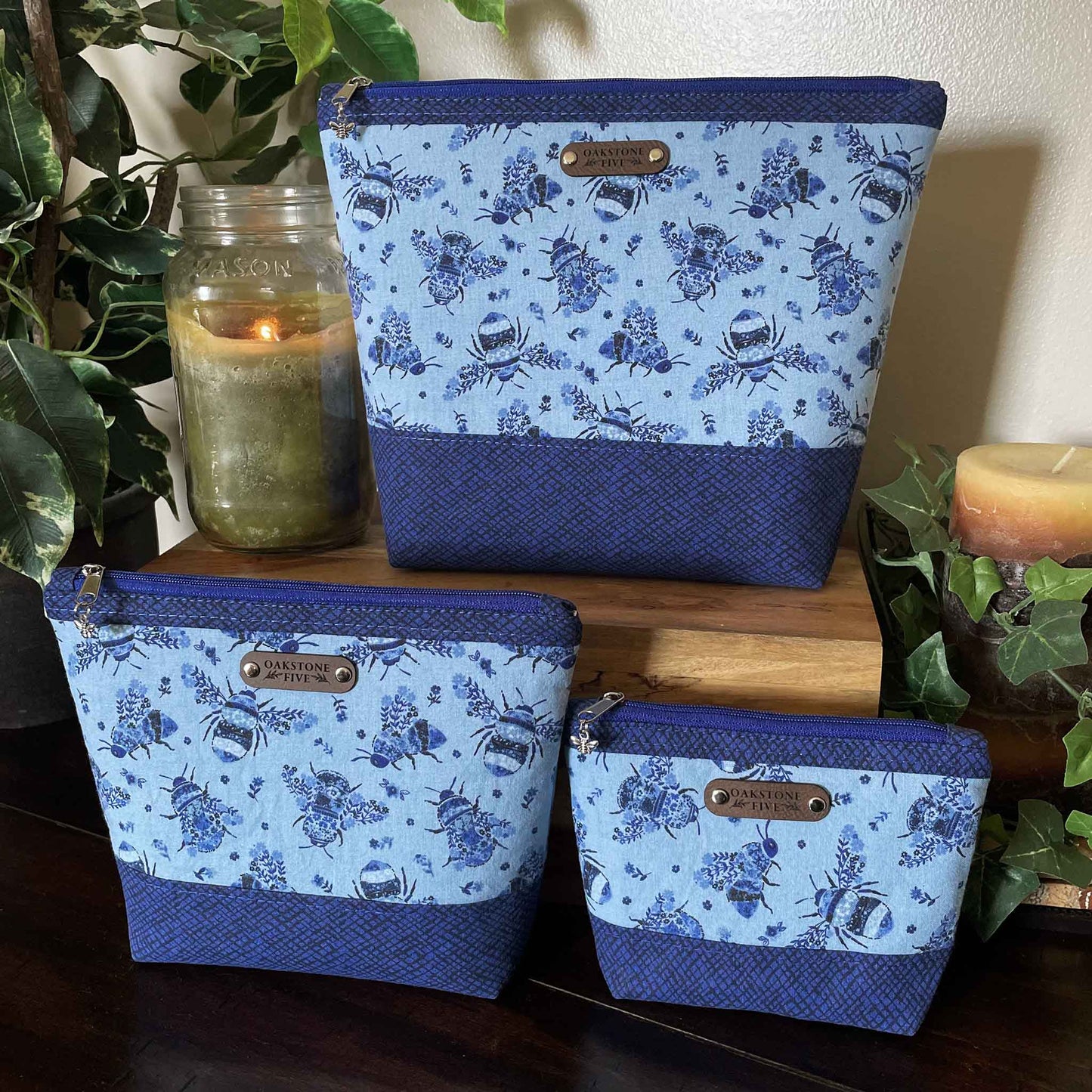 Set of 3 blue floral bumble bee patterned zipper pouches, sitting on a wooden table with a candle and plants.