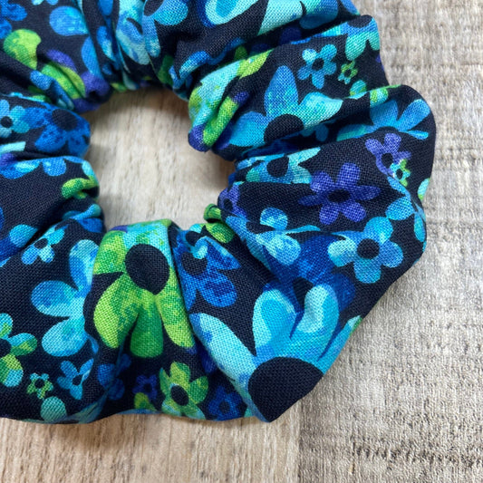A close-up of a cute, blue and green daisy floral scrunchie laying on a wooden table.