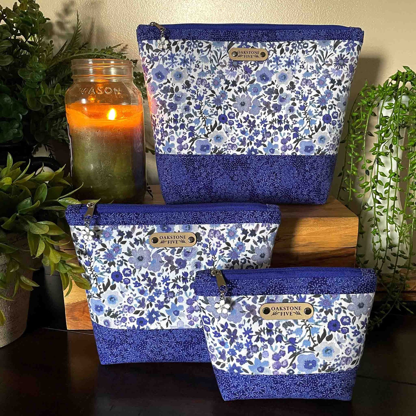 Set of three blue floral-patterned zipper pouches with "Oakstone Five" branding on a wooden surface with plants and a candle in the background.