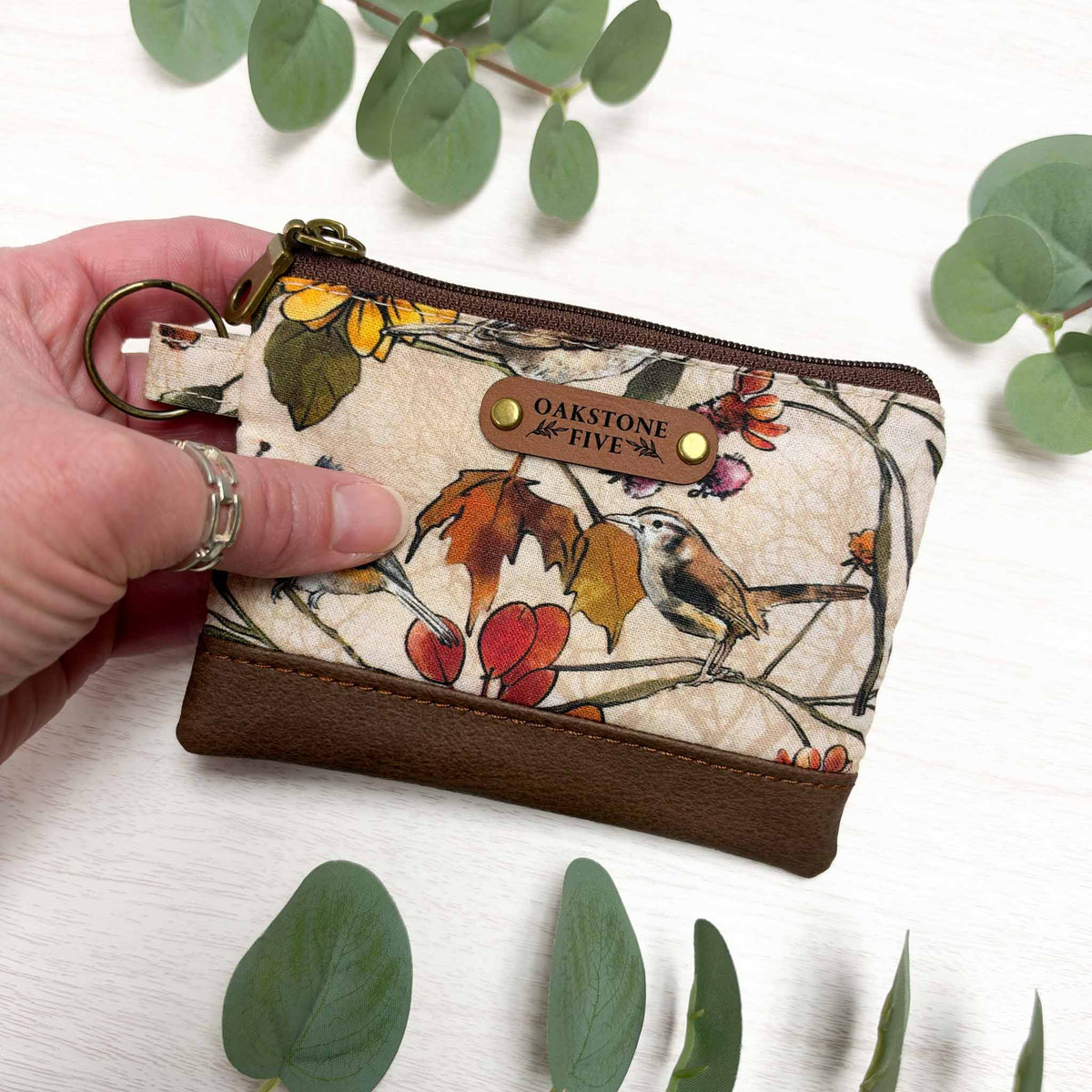A hand holding a small zipper pouch with a floral and bird print. The pouch has a brown zipper and faux-leather bottom, with brass hardware. 