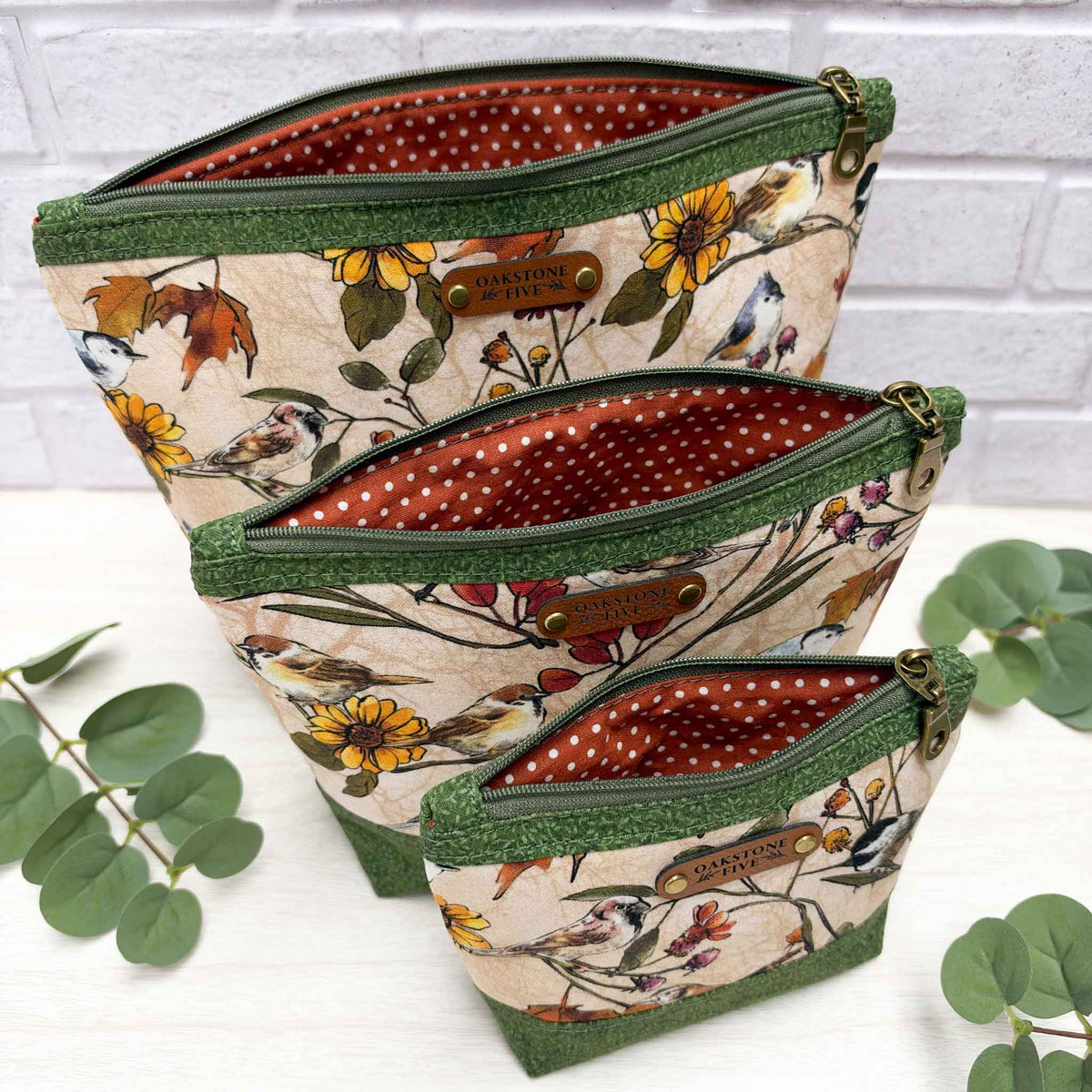 Three fabric zip pouches of different sizes arranged in a nested stack on a white surface with eucalyptus leaves. The pouches feature a beige background with a detailed bird and floral print, green vines trim, and a burnt orange polka dot lining.