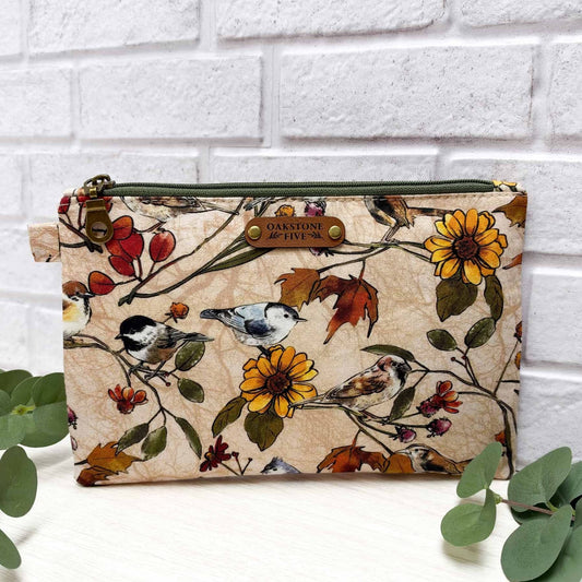 A zippered pouch with a nature print featuring birds, sunflowers, and leaves on a beige background, styled with eucalyptus leaves in front of a white brick wall.