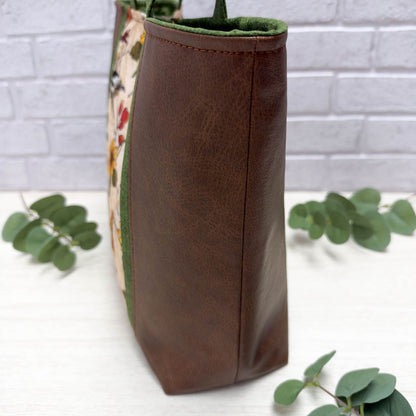 A close-up of the side of a brown faux-leather tote bag with a floral and bird printed fabric panel on the side, green trim, and short, green handles, shown standing on a white surface with eucalyptus leaves and a white brick wall background.