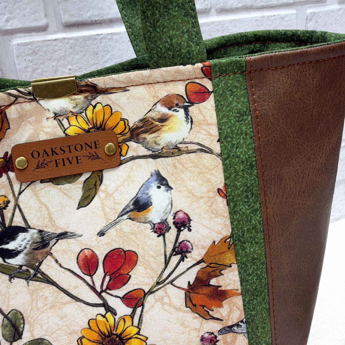 Close-up of a fabric tote bag with a bird and floral print on a cream background, green patterned fabric trim, a brown faux-leather side panel, and a faux leather brand tag reading Oakstone Five.