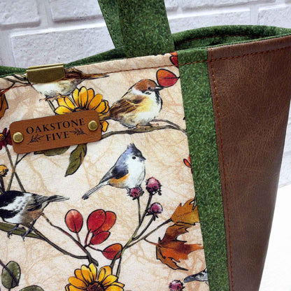 Close-up of a fabric tote bag with a bird and floral print on a cream background, green patterned fabric trim, a brown faux-leather side panel, and a faux leather brand tag reading Oakstone Five.