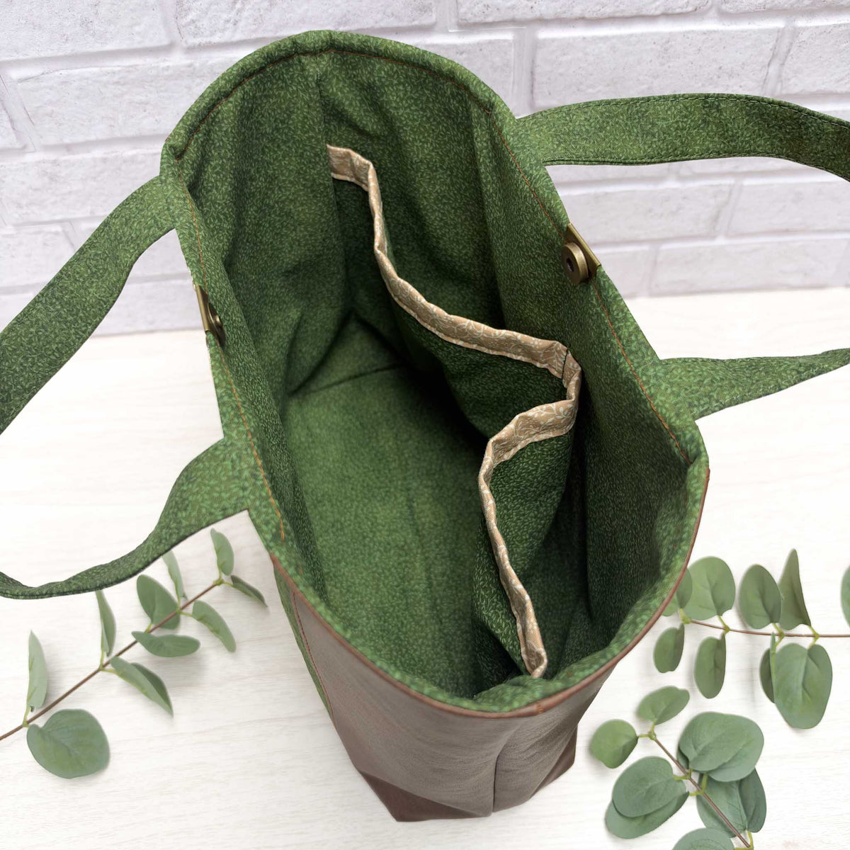 An open green vines, nature-inspired tote bag with interior slip pockets, and short, hand carry handles, photographed on a white surface with eucalyptus leaves and a white brick wall background.