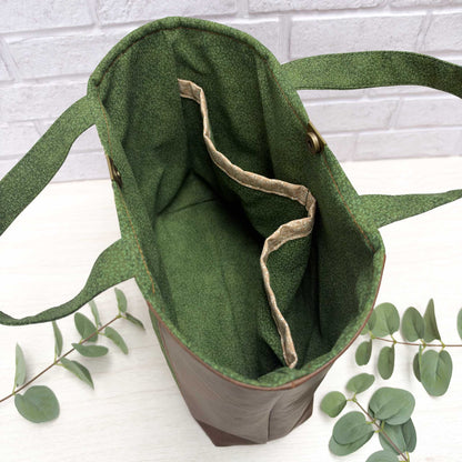 An open green vines, nature-inspired tote bag with interior slip pockets, and short, hand carry handles, photographed on a white surface with eucalyptus leaves and a white brick wall background.