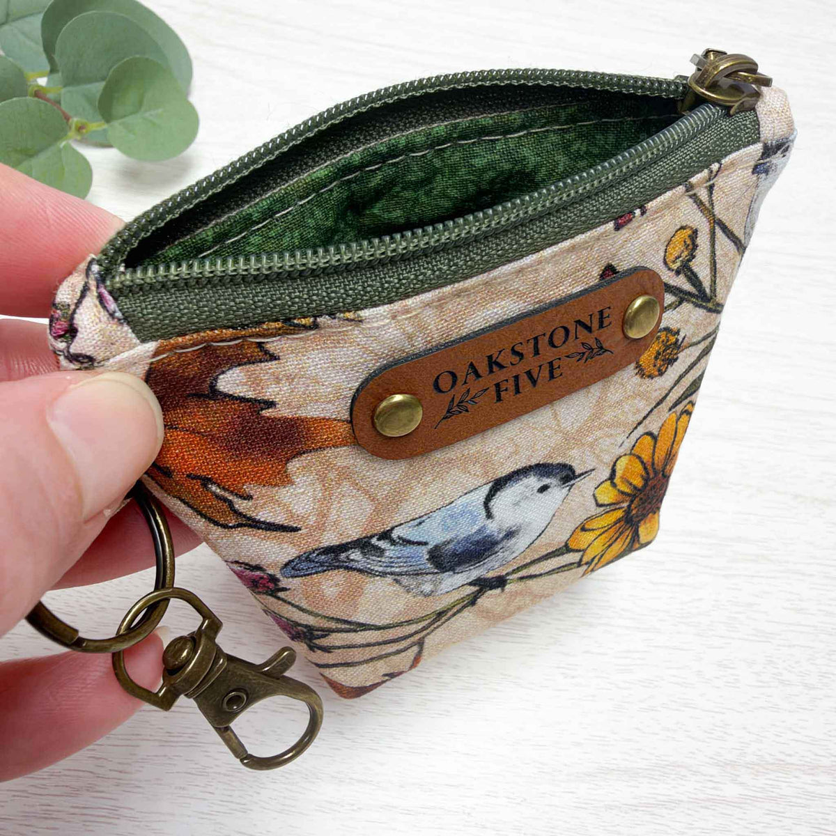 A hand holds an open, small zippered coin pouch with a floral and bird print. The pouch has an olive green zipper, a green vines interior liner, and brass hardware, a key ring and snap hook.