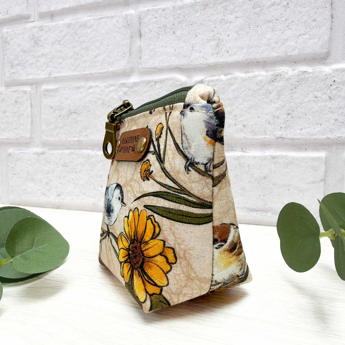 A side view of a small, cotton fabric zippered pouch with a cream background with a bird and floral print. Photographed on a white surface, with green leaves and a white brick backround.