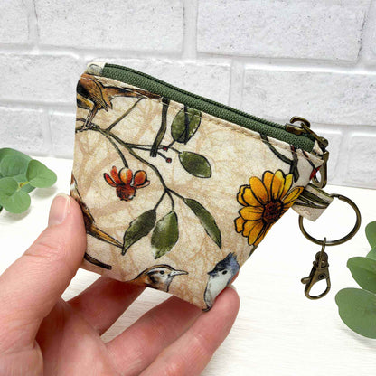 A hand holding a small zippered fabric coin purse with a bird and flower print, featuring a green zipper and brass keychain clasp, set against a white brick wall background.