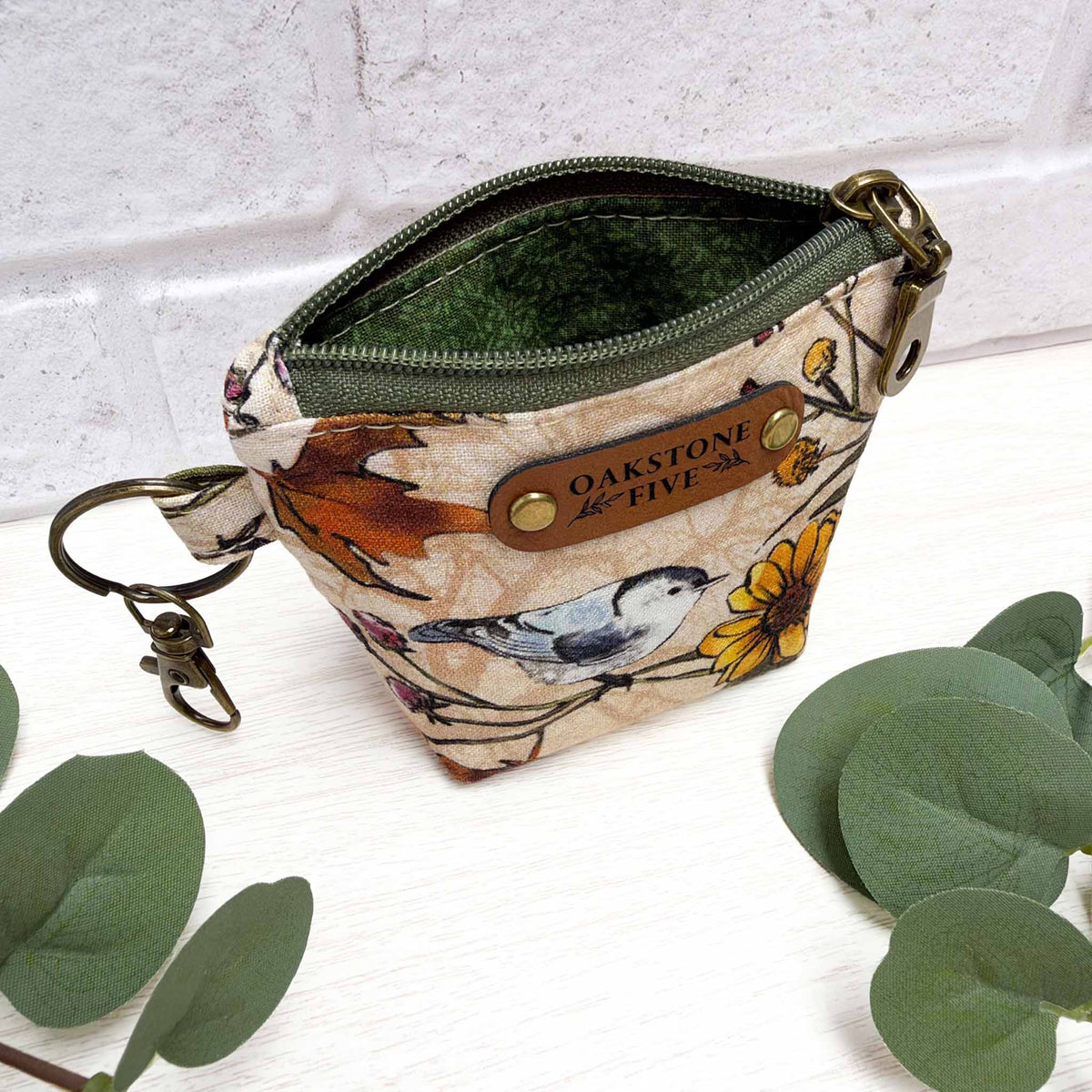 A mini keychain zip pouch with a bird and flower print exterior featuring a brown faux leather brand tag, and brass hardware, with a green vines liner, photographed on a white surface with decorative green leaves and a white brick wall background.