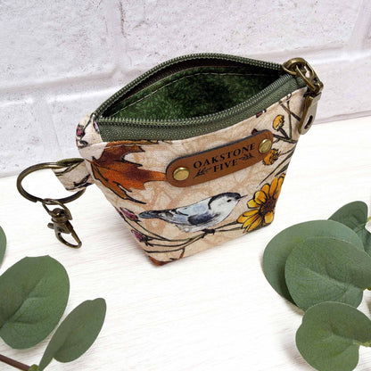A mini keychain zip pouch with a bird and flower print exterior featuring a brown faux leather brand tag, and brass hardware, with a green vines liner, photographed on a white surface with decorative green leaves and a white brick wall background.