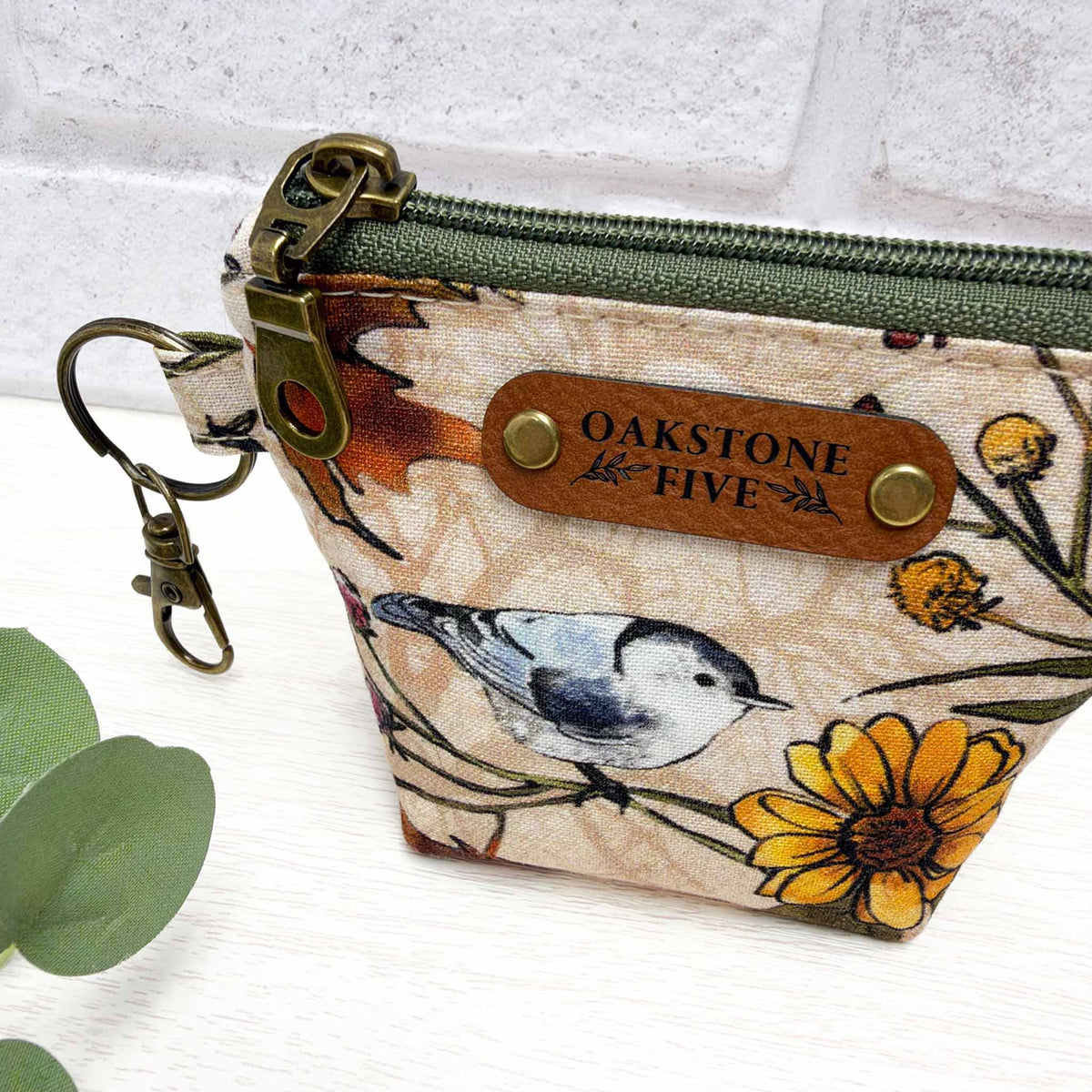 A small keychain zip pouch with a bird and flower print, featuring a green zipper, brass hardware, and a faux leather brand tag, photographed on a light surface with a white brick background.