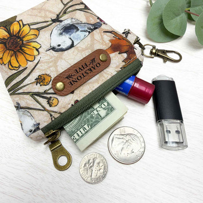 A small printed keychain zip pouch with a bird and flower pattern, olive green zipper, brass-tone hardware, and a brown faux leather "Oakstone Five" brand tag. The pouch is shown open with a dollar bill, chapstick, USB flash drive, and coins beside it on a white surface with eucalyptus leaves in the corner.