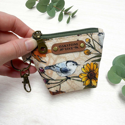 A hand holds a mini zip keychain pouch with a bird and flower print. The pouch has a brown faux leather "Oakstone Five" brand tag and an antique brass keyring clasp.