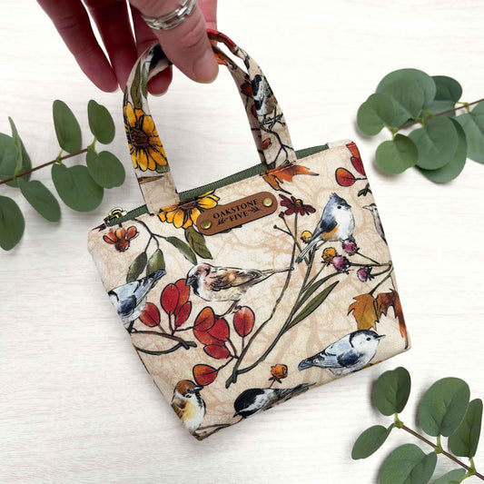 A hand holds the top handle of a mini zippered tote bag pouch with a bird and floral print. The bag has a beige background with illustrated birds, red berries, yellow flowers, and a brown faux leather "Oakstone Five" brand tag.