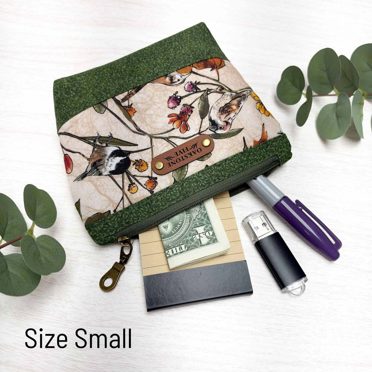 A small zipper pouch with a bird and floral pattern and green vines accent fabric, shown on a white surface with eucalyptus leaves. The zip pouch is styled with cash, a notepad, a marker, and a USB drive.