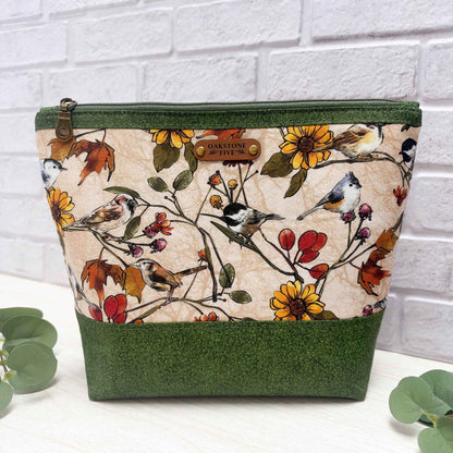A large sized, organizational zipper bag with a green vines fabric trim and base, featuring a central printed panel of birds and flowers on a beige background, with a brown faux leather Oakstone Five brand patch on the front.