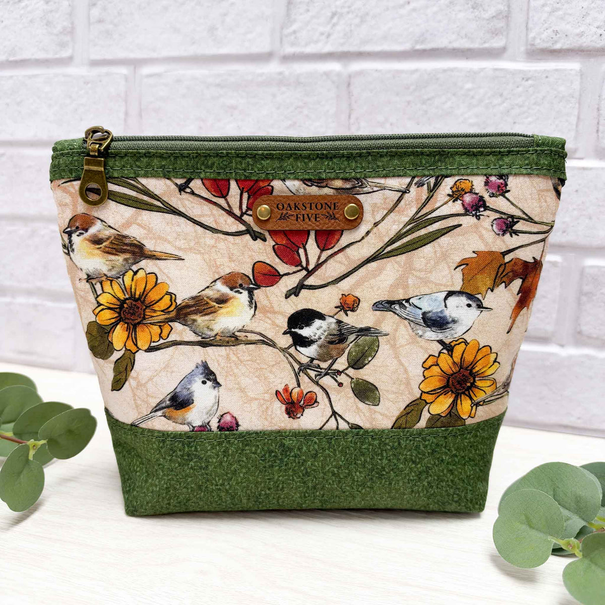 A medium sized zippered pouch with a green vines fabric trim and base, featuring a central printed panel of birds and flowers on a beige background, with a brown faux leather Oakstone Five brand patch on the front.